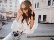 Glad young woman with black wavy hair laughing on the street. Outdoor shot of cute girl in sunglasses posing with camera.