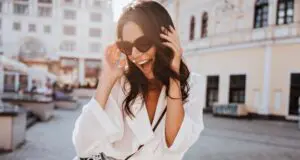 Glad young woman with black wavy hair laughing on the street. Outdoor shot of cute girl in sunglasses posing with camera.