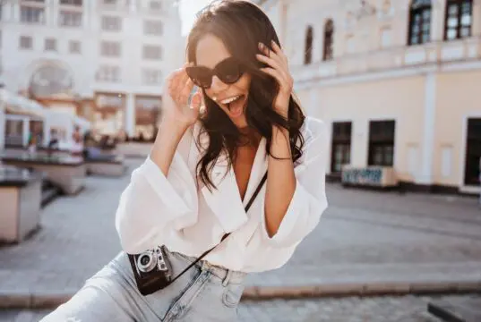 Glad young woman with black wavy hair laughing on the street. Outdoor shot of cute girl in sunglasses posing with camera.