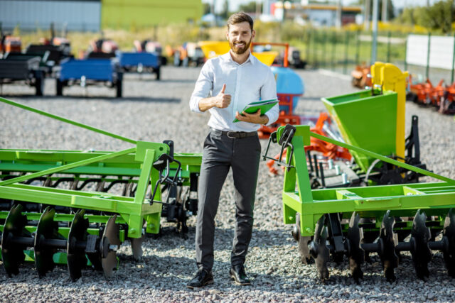 Portrait of a handsome salesman standing near the plow at the outdoor ground of the shop with new agricultural machinery