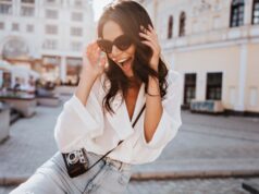 Glad young woman with black wavy hair laughing on the street. Outdoor shot of cute girl in sunglasses posing with camera.